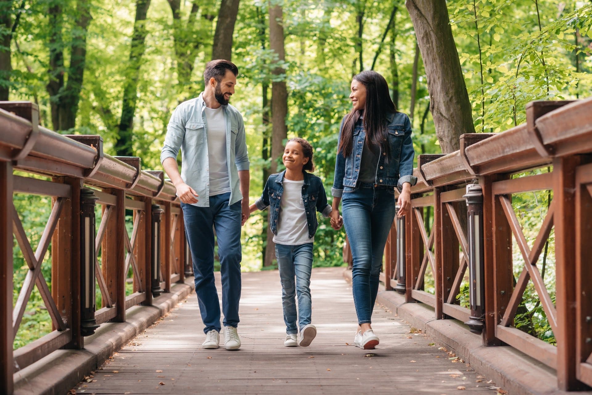 mom, dad and daughter walking on a small bridge in woods